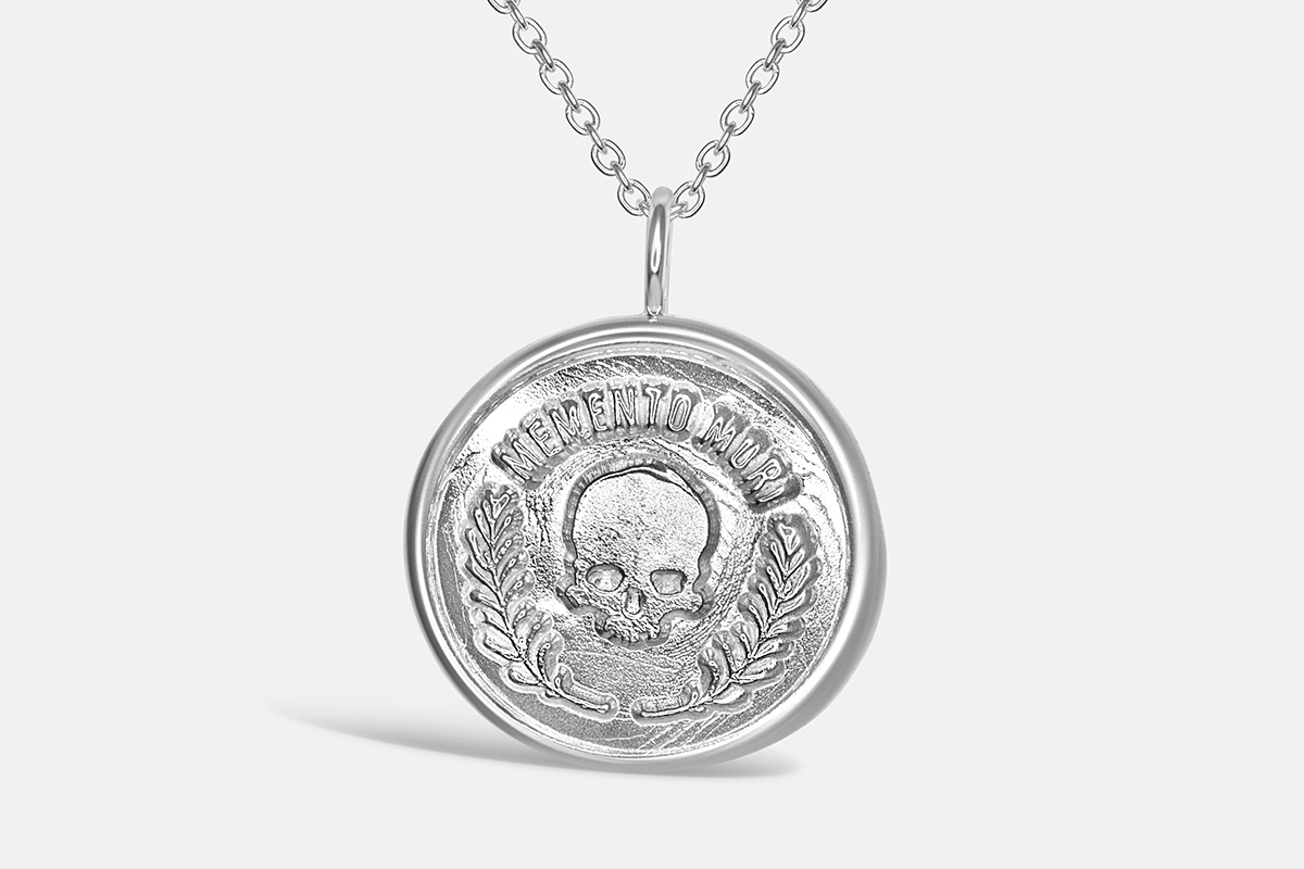 Large silver memento mori pendant on a whilte background. A skull at the centre topped by the words Memento Mori and surrounded by rosemary leaves underneath.