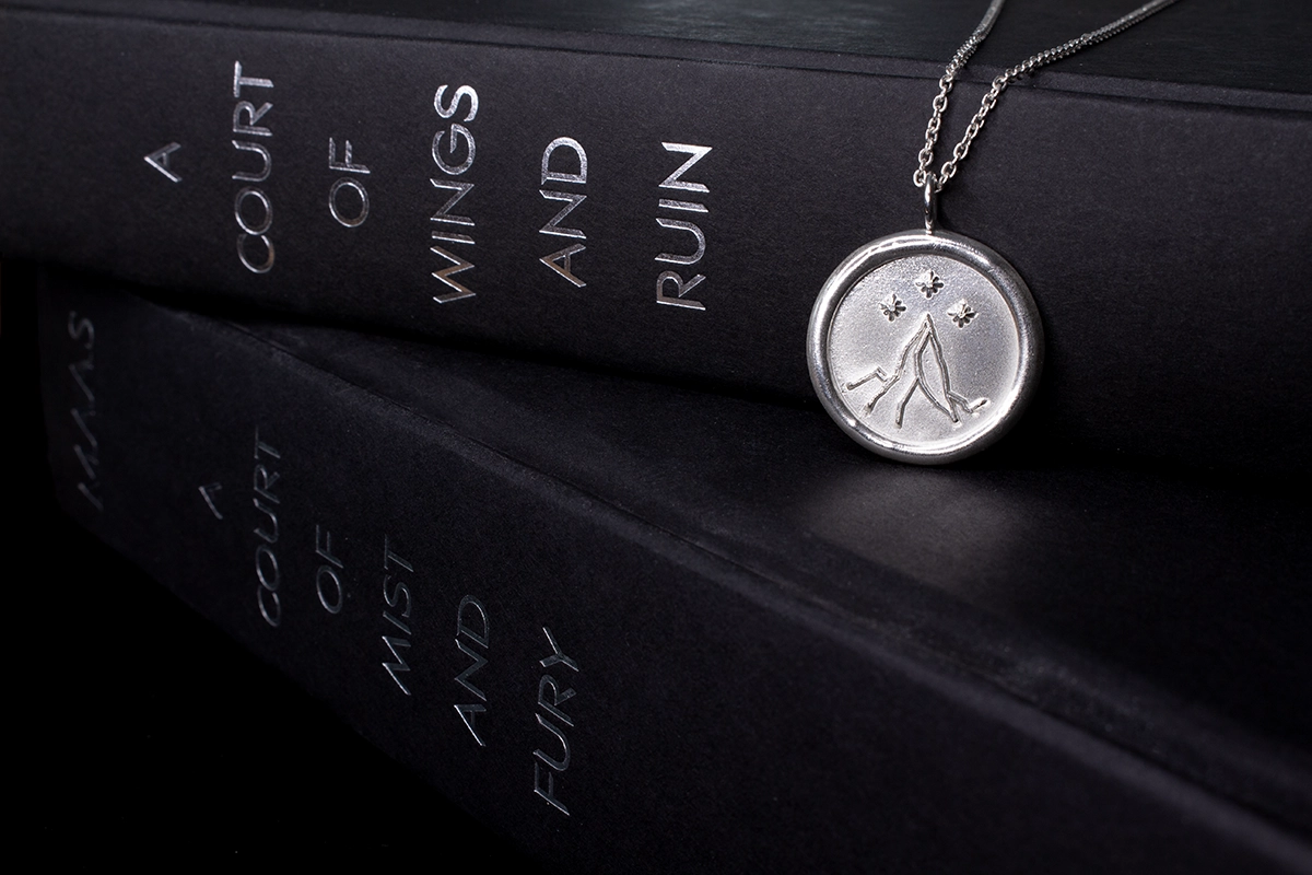 Two black hardcover books with silver text "A court of Mist and Fury" and "A Court of Wings and Ruin" with a silver night court pendant draped over the stack of books.