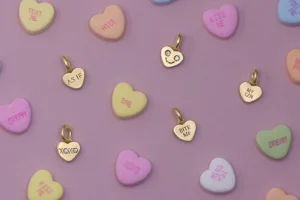 Solid gold conversation heart pendants photographed on pink paper covered with candy hearts.